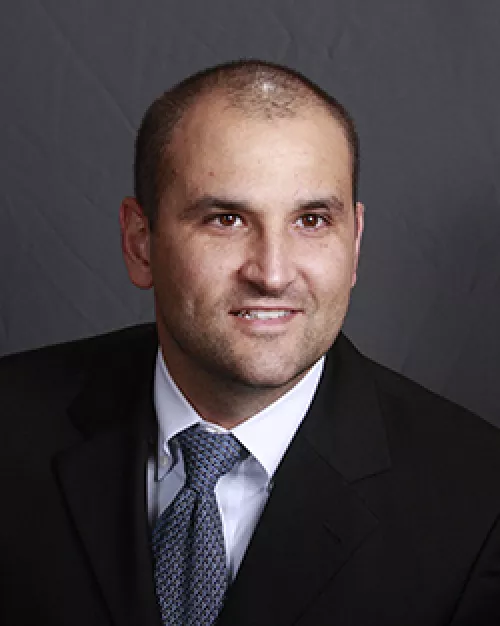 Portrait of a doctor in a suit smiling against a gray background.