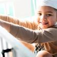 A toddler with gauze bandages around his head stands in his crib and smiles at the camera.