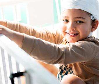 A toddler with gauze bandages around his head stands in his crib and smiles at the camera.
